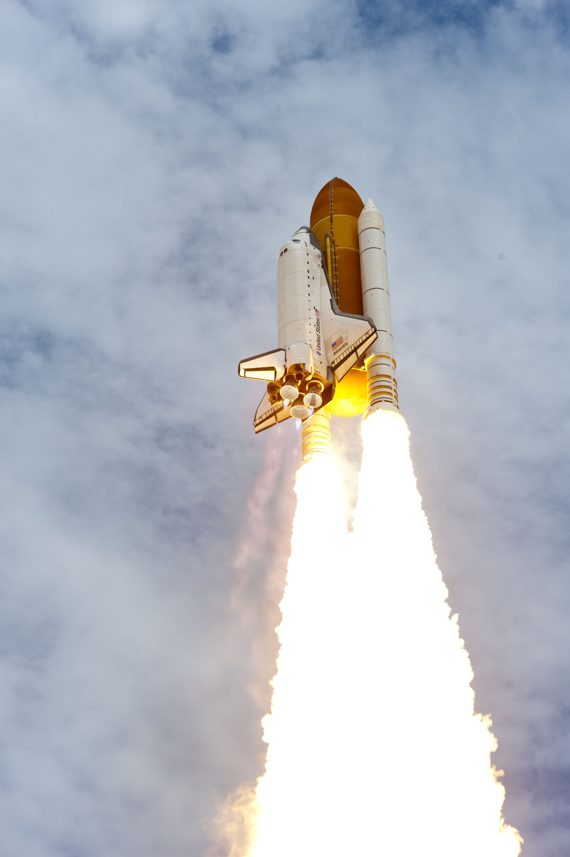 Space Shuttle Atlantis lifting off with glowing exhaust against a cloudy sky